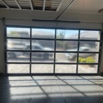 Glass-panel garage door with metal frame, sunlight casting shadows inside, vehicles and houses visible outside.